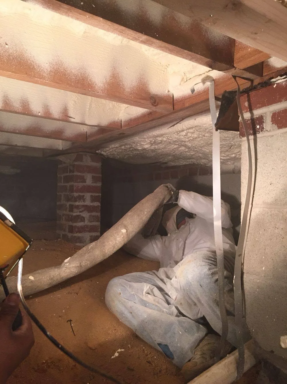 Insulation contractor in a white protective suit applying spray foam in a tight crawl space under floor joists